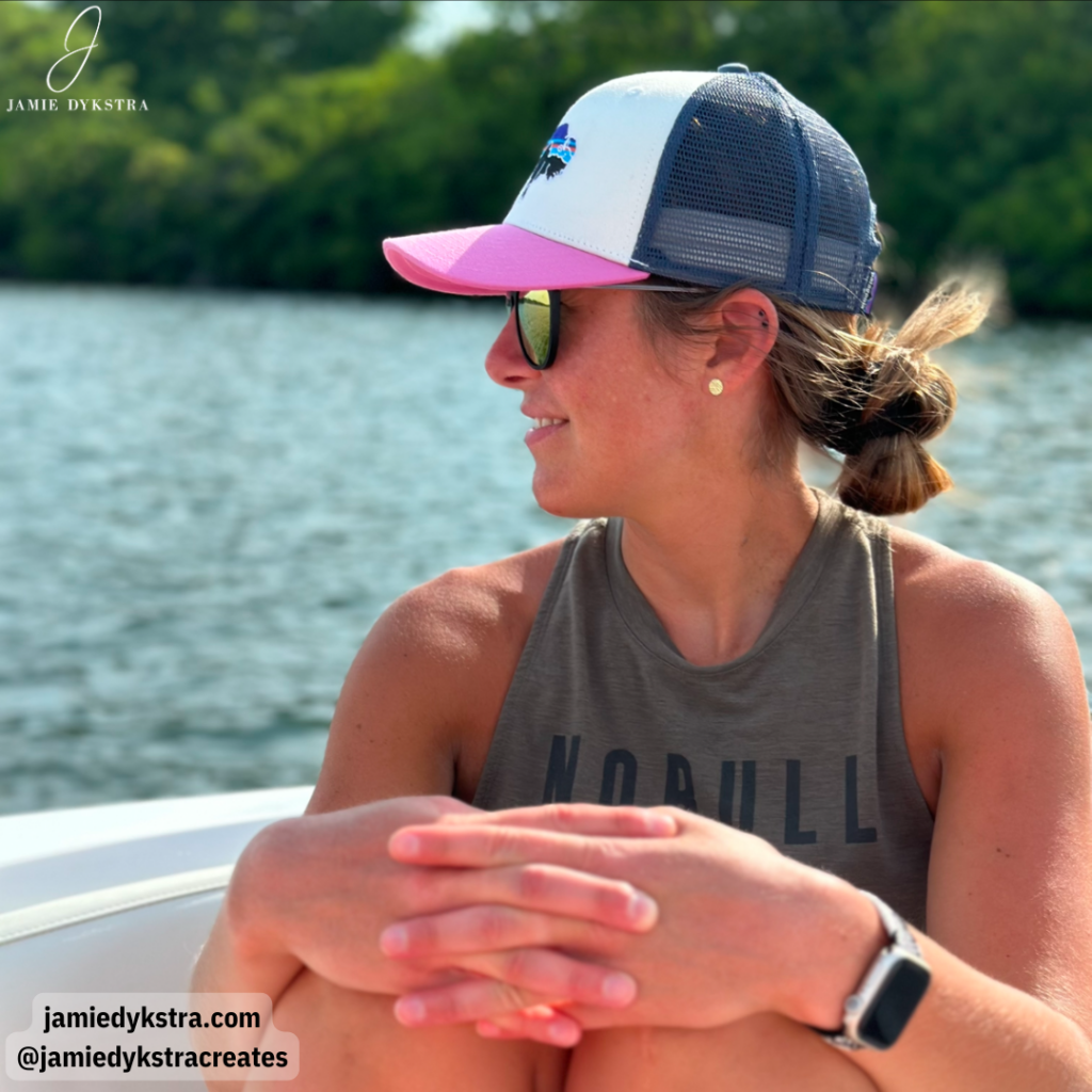 A picture of me in a pink and navy baseball hat with a dark green tank top on a boat.