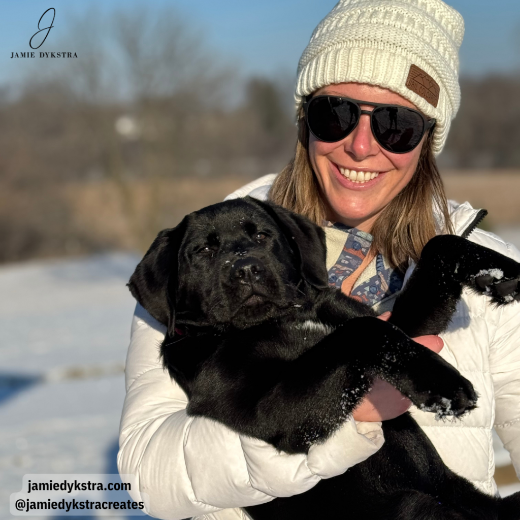 A picture of me with a white knit hat and black sunglasses standing outside on a sunny day with snow in the background holding my Black Lab puppy.