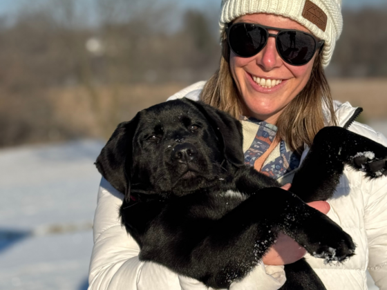 A picture of me with a white knit hat and black sunglasses standing outside on a sunny day with snow in the background holding my Black Lab puppy.