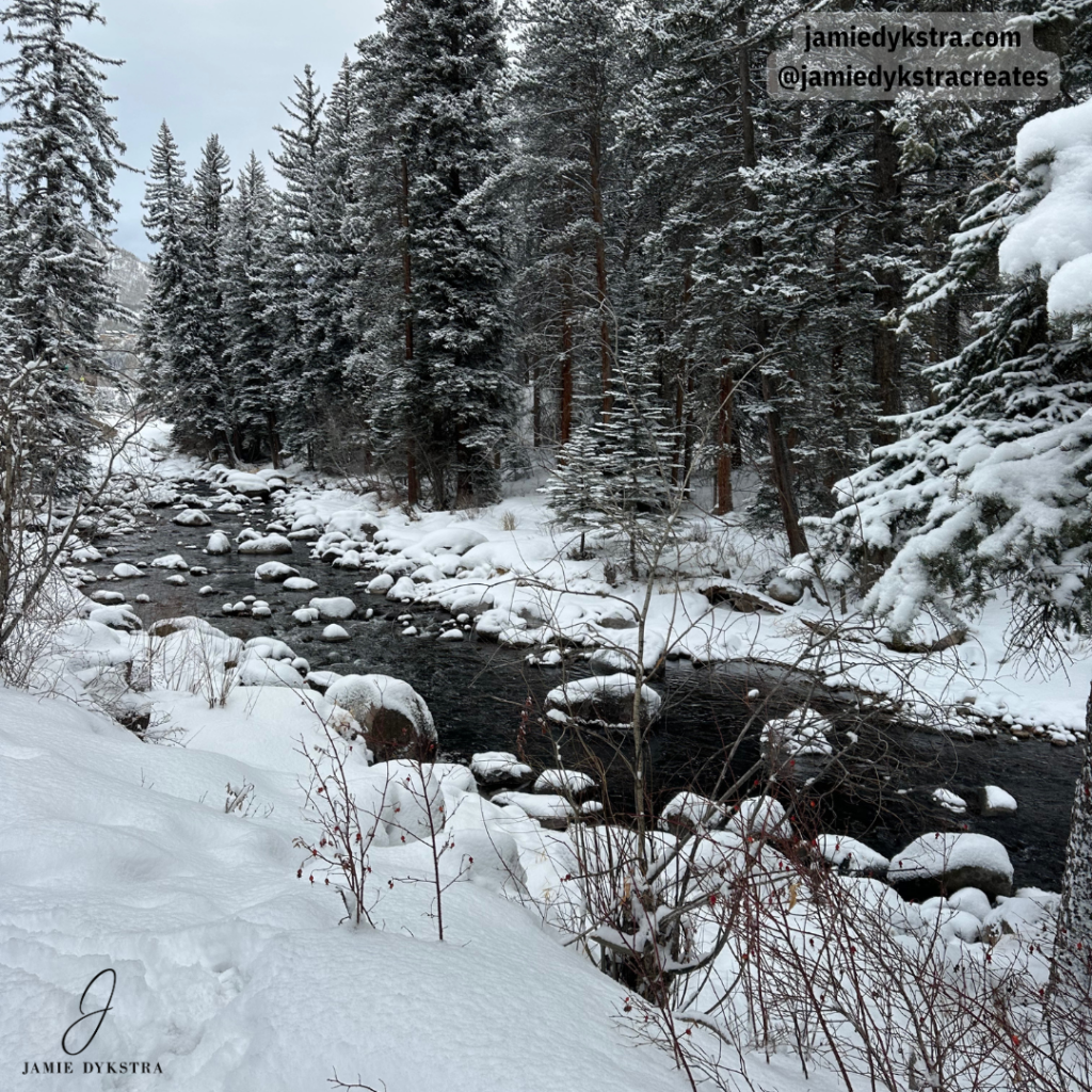 A picture of a walking trail along a creek with a fresh coat of powdery snow and evergreen trees towering over the creek at the base of a mountain.