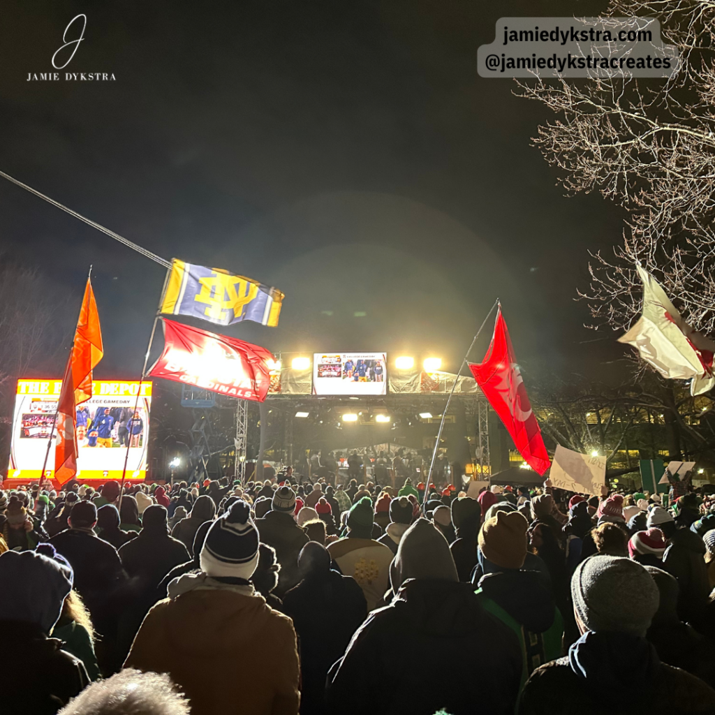 A photo in a crowd of people at the set of Collage Gameday at night with flags of different colleges hoisted in the air.