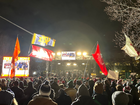 A photo in a crowd of people at the set of Collage Gameday at night with flags of different colleges hoisted in the air.