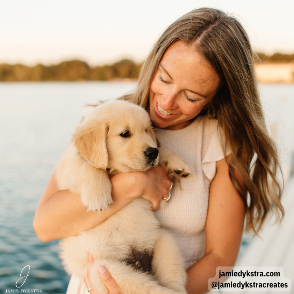 A picture of me on a dock over a lake with my hair down in a cream dress holding a golden retriever puppy.
