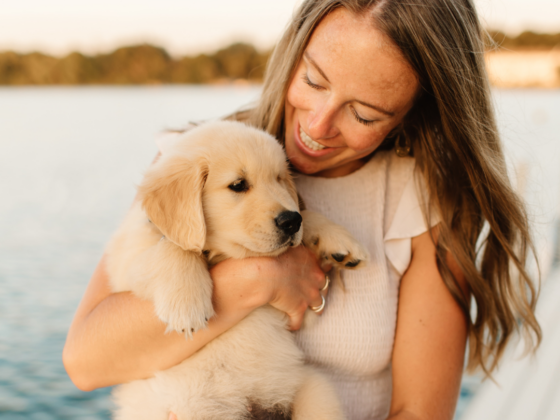 A picture of me on a dock over a lake with my hair down in a cream dress holding a golden retriever puppy.