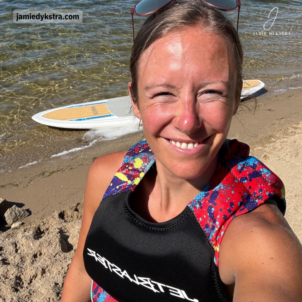 A selfie of me in a life jacket with sand and a paddleboard in the water behind me.
