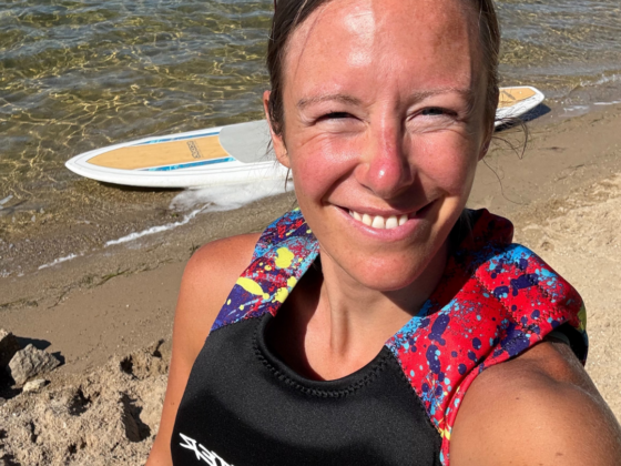 A selfie of me in a life jacket with sand and a paddleboard in the water behind me.