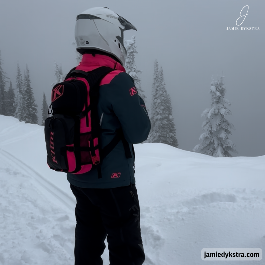 A picture of me standing at the top of a mountain with snow on the ground and fog all around. I have one a white helmet, black and pink jacket, black snowmobile pants, and a black and pink backpack.