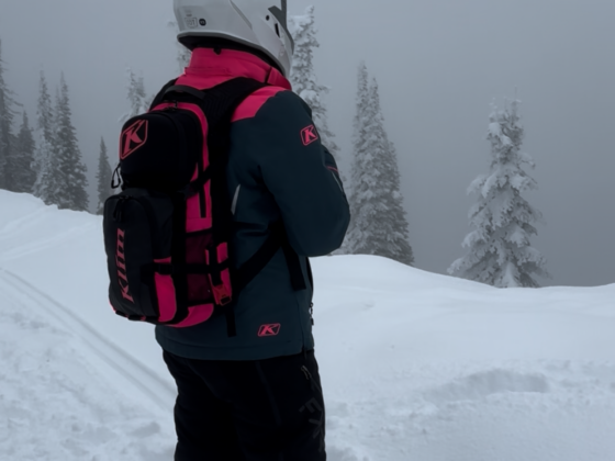 A picture of me standing at the top of a mountain with snow on the ground and fog all around. I have one a white helmet, black and pink jacket, black snowmobile pants, and a black and pink backpack.