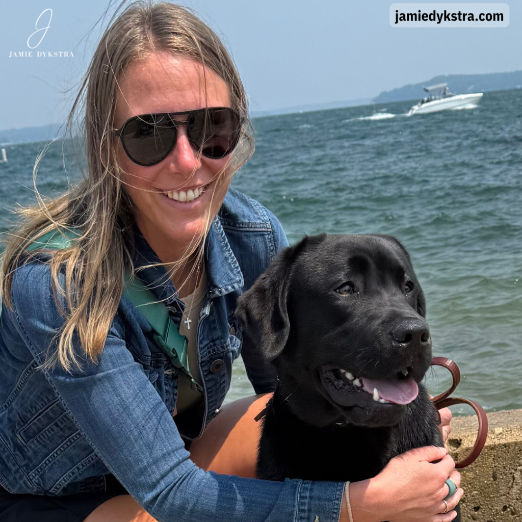 A picture of me with me kneeling down with my arms around my black lab in front of a big lake with boats on it.
