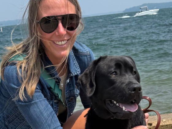A picture of me with me kneeling down with my arms around my black lab in front of a big lake with boats on it.