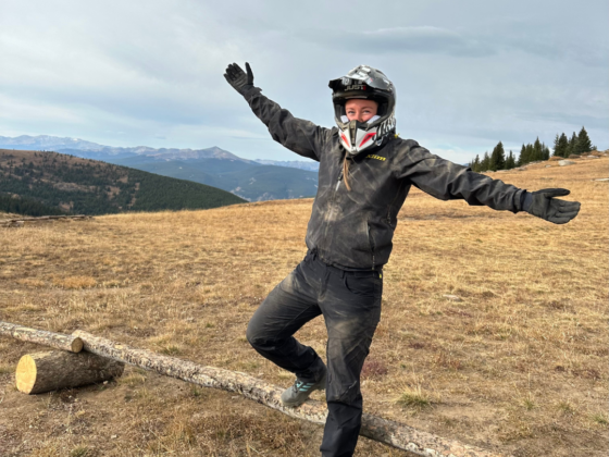 A picture of me in a helmet and dusty riding gear with arms spread out at the top of a mountain after riding side by sides up the trail.