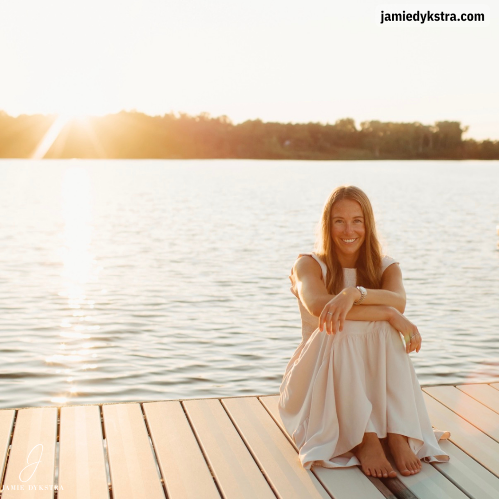 I picture of me in a long cream dress, sitting with my arms around my knees, on a pier with a lake behind me as the sun begins to set.