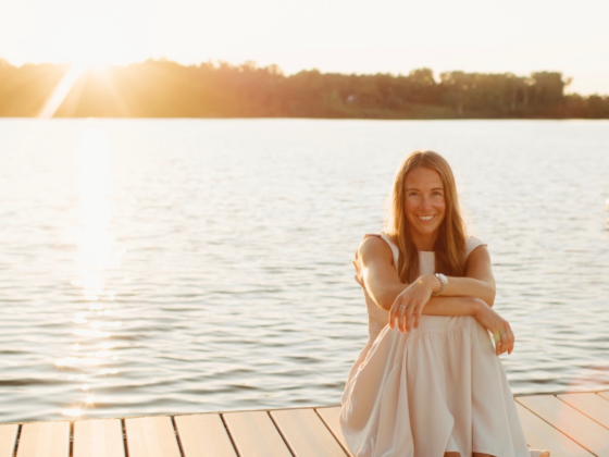 I picture of me in a long cream dress, sitting with my arms around my knees, on a pier with a lake behind me as the sun begins to set.