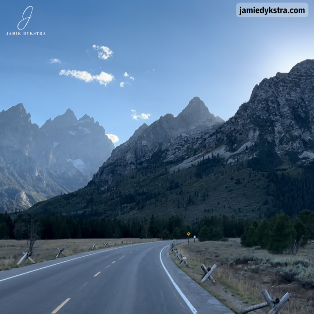 An image of a road headed toward the Grand Teton mountains with sun behind the mountains.