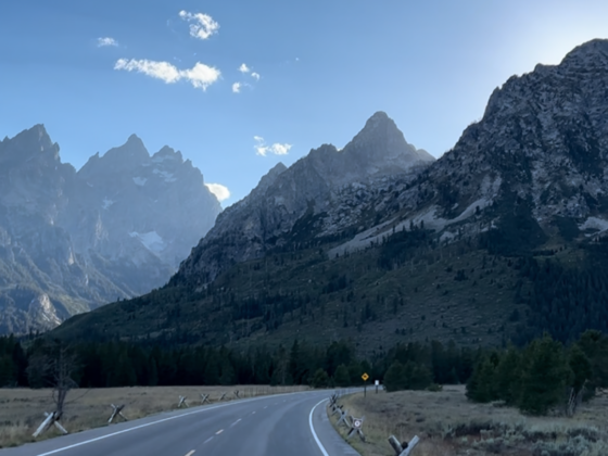 An image of a road headed toward the Grand Teton mountains with sun behind the mountains.
