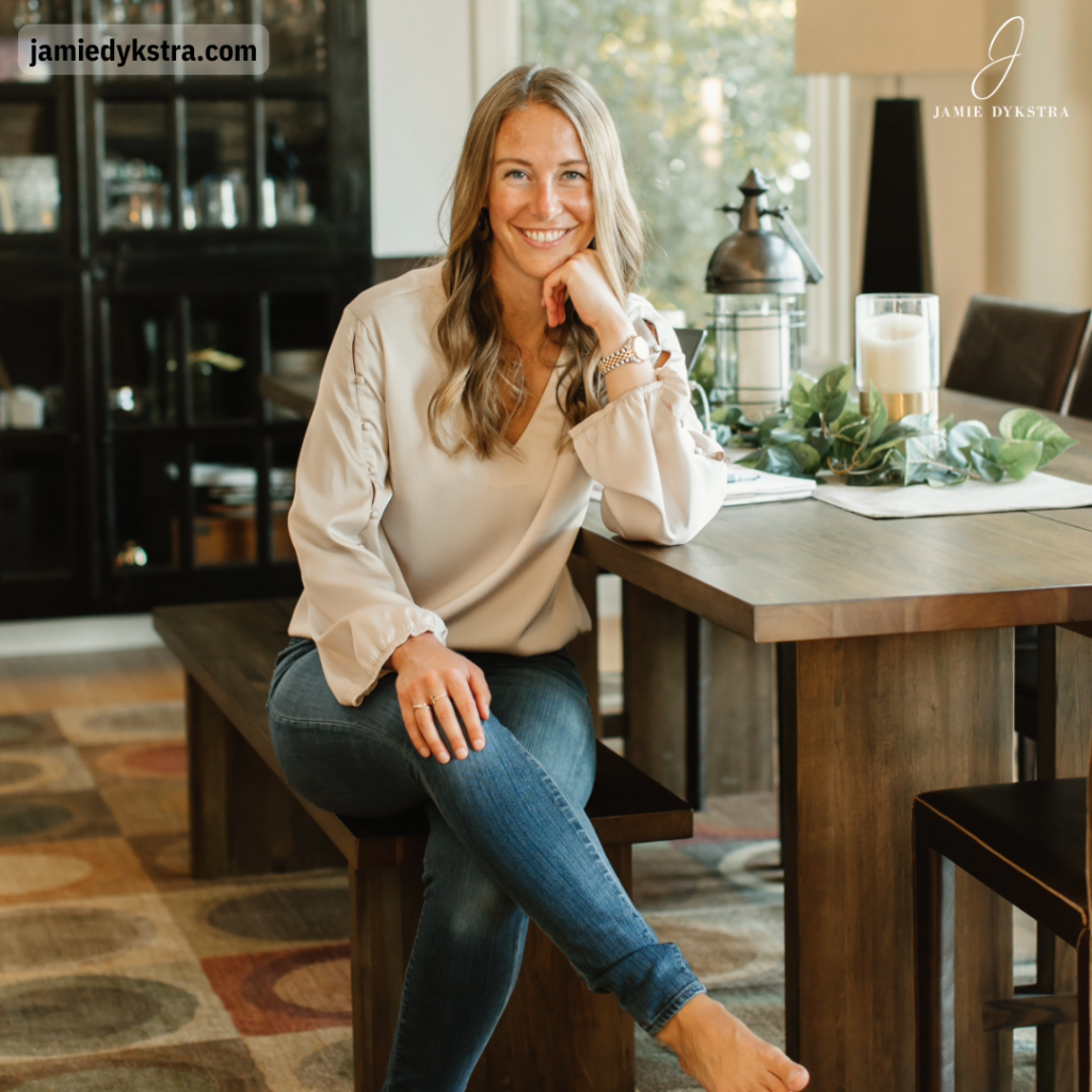A photo of me sitting at a dining room table in a cream top and jeans with my legs crossed and my chin resting on my hand.