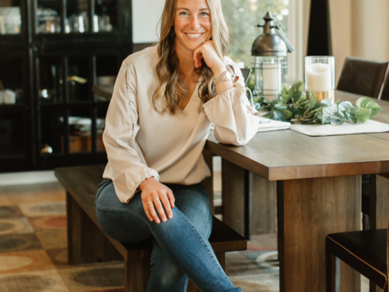 A photo of me sitting at a dining room table in a cream top and jeans with my legs crossed and my chin resting on my hand.