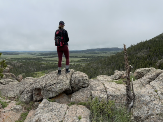 A photo of me standing on some large rocks on a mountain with my back to the camera overlooking a valley and foothills.