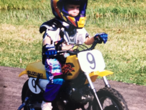 A photo of a young me on a yellow dirt bike with training wheels and a yellow and purple outfit and helmet.