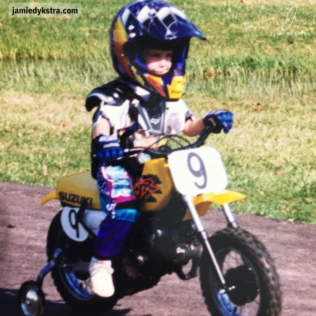 A photo of a young me on a yellow dirt bike with training wheels and a yellow and purple outfit and helmet.