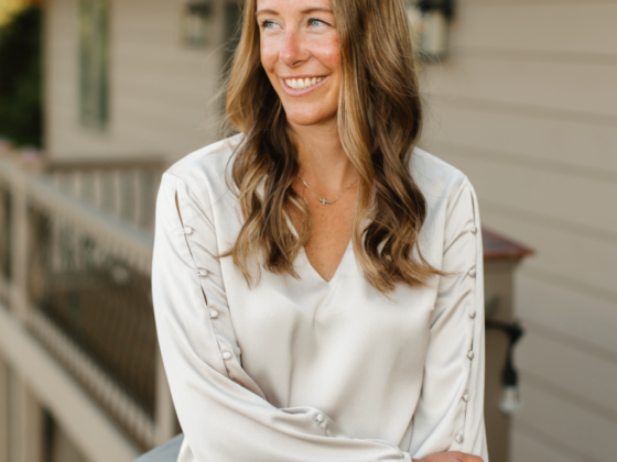 A photo of Jamie Dykstra standing on a deck with one arm over another in a cream silk top.