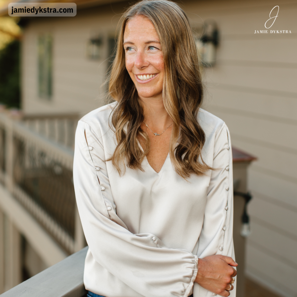 A photo of Jamie Dykstra standing on a deck with one arm over another in a cream silk top.