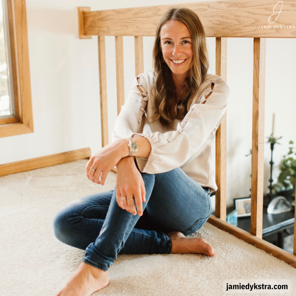 A photo of me sitting on the ground with one leg crossed under the other, leaning against a railing, in a cream silk shirt and jeans.