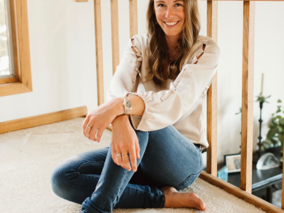 A photo of me sitting on the ground with one leg crossed under the other, leaning against a railing, in a cream silk shirt and jeans.