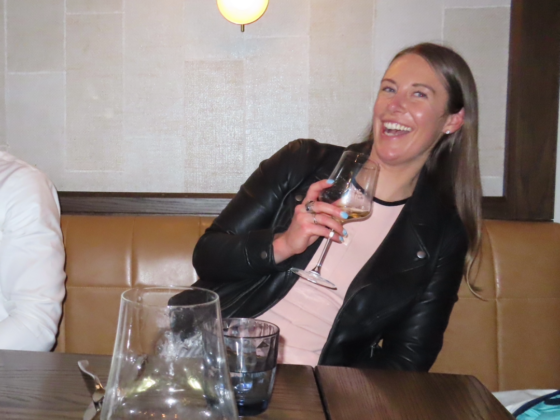 A photo of my leaning out of a picture holding a glass of wine in a light pink dress with a black leather jacket.